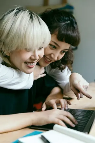 positive_smiling_asian_girlfriends_hugging_and_looking_at_laptop_screen_while_working_on_homework_together_at_table_with_opened_notebook.webp