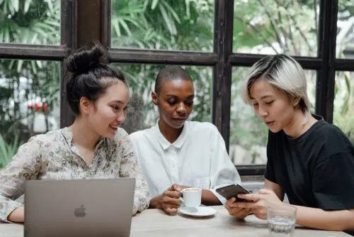 young_asian_and_african_american_women_checking_posts_on_social_media_while_sitting_in_cozy_cafe_with_coffee_and_laptop_near_large_window_with_exotic_green_garden_outside.webp
